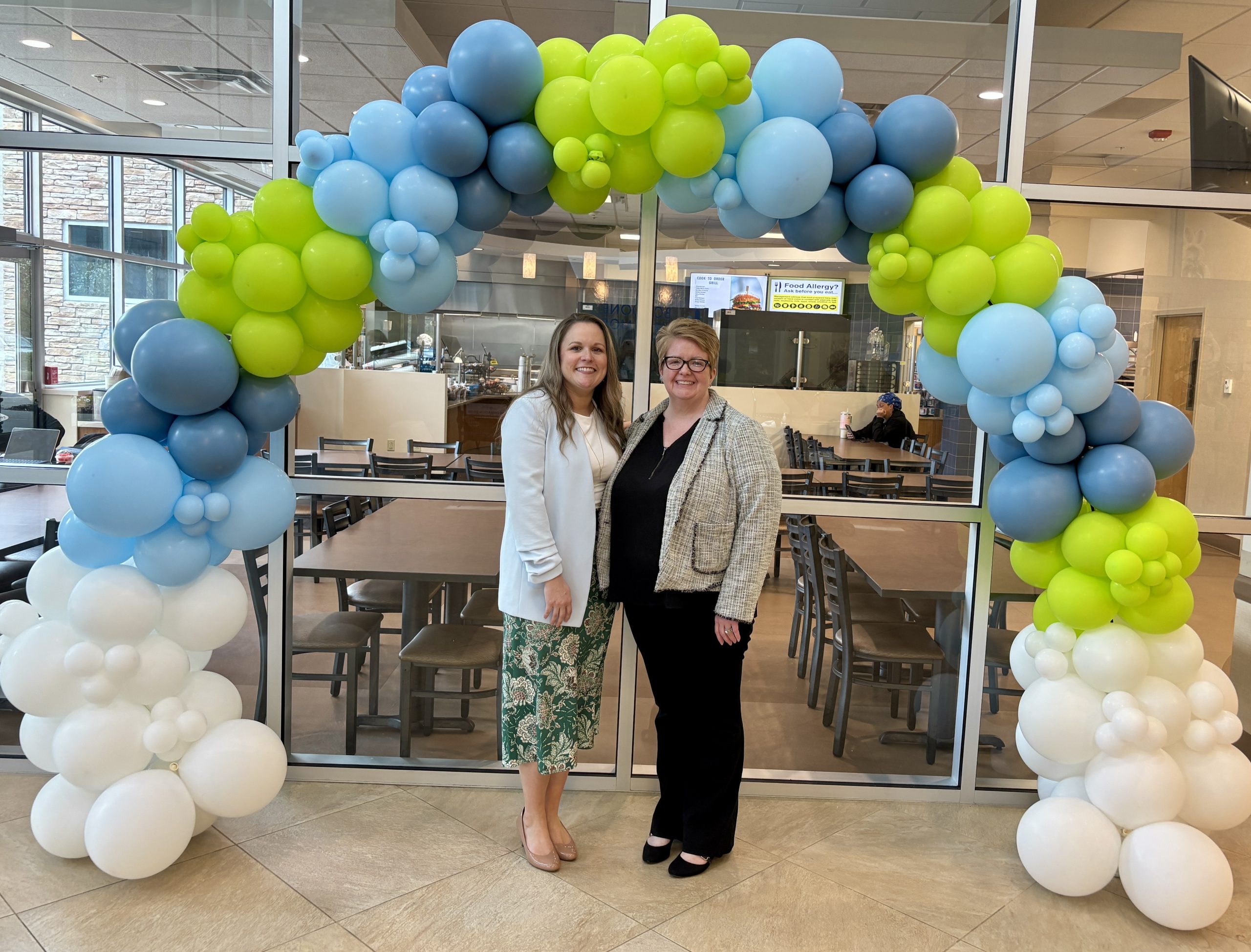 Two women stand beneath a blue and green balloon arch outside the BMH hospital cafeteria