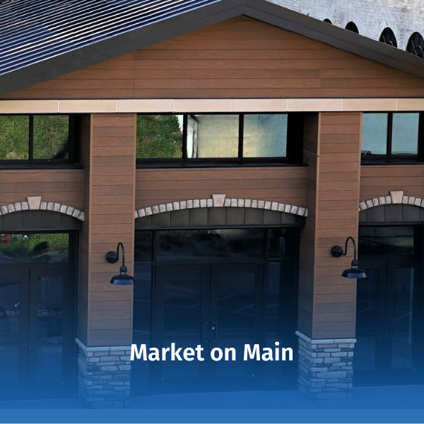 Front entrance of the Market on Main building, featuring a modern design with wood siding, stone columns, large windows, and exterior lighting. The image represents a community gathering place that offers local products, events, and activities. Text overlay reads “Market on Main.”